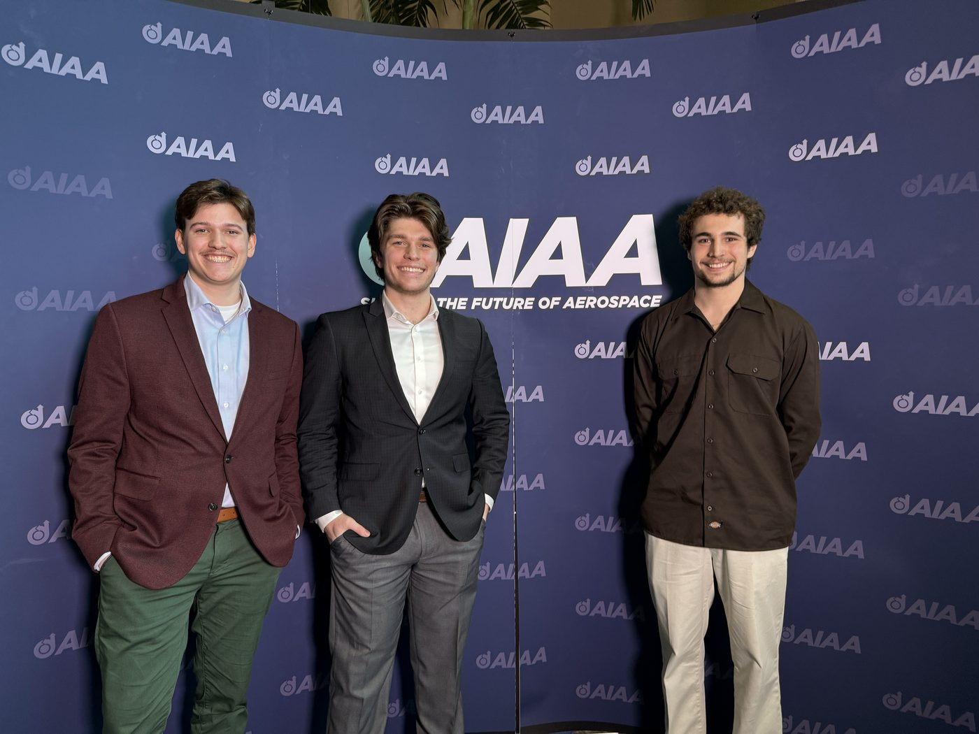 Three researchers standing in front of an AIAA branded backdrop at a conference.