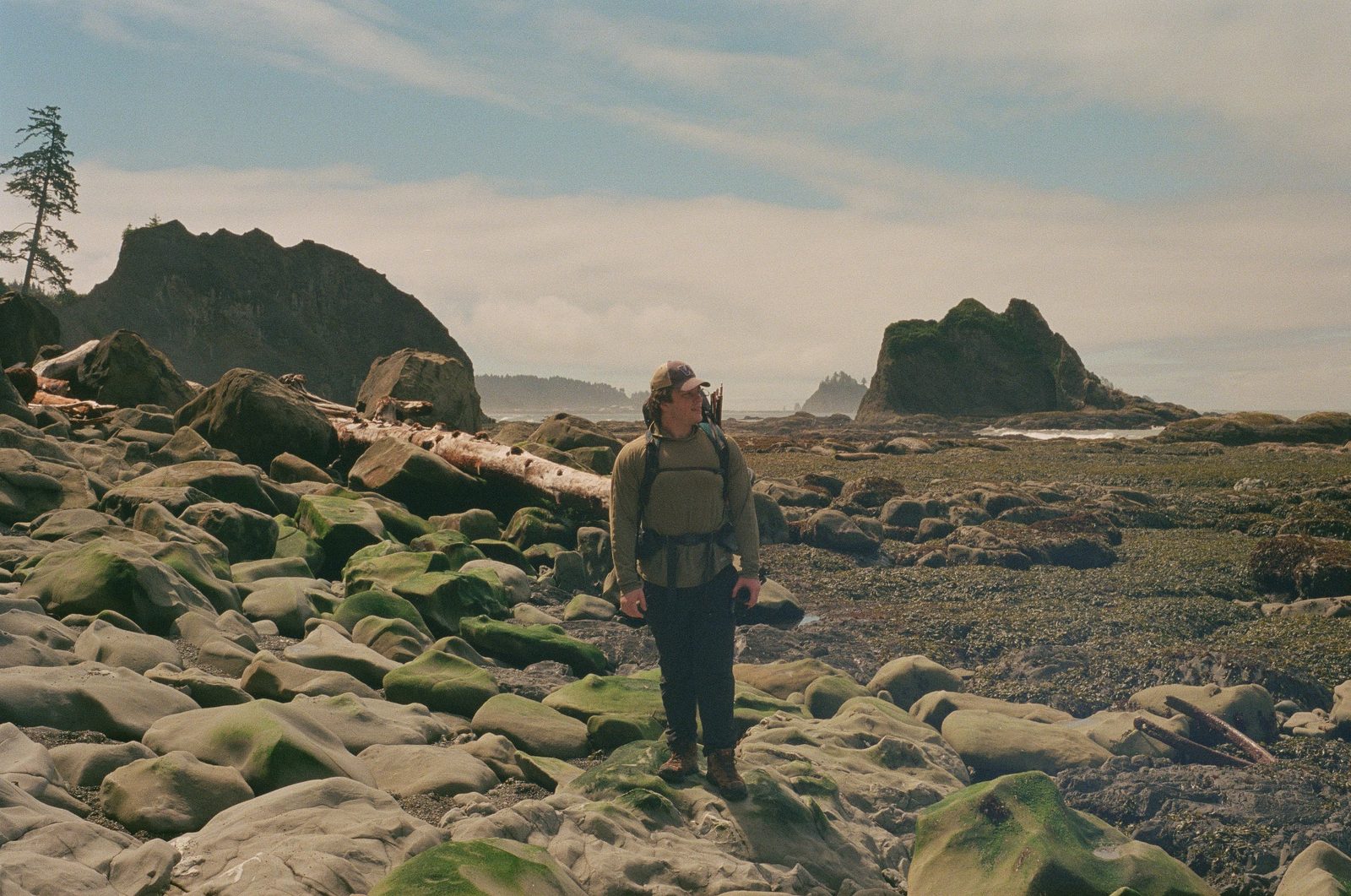 A backpacker standing among algae-covered boulders on a rocky coastline with sea stacks behind.