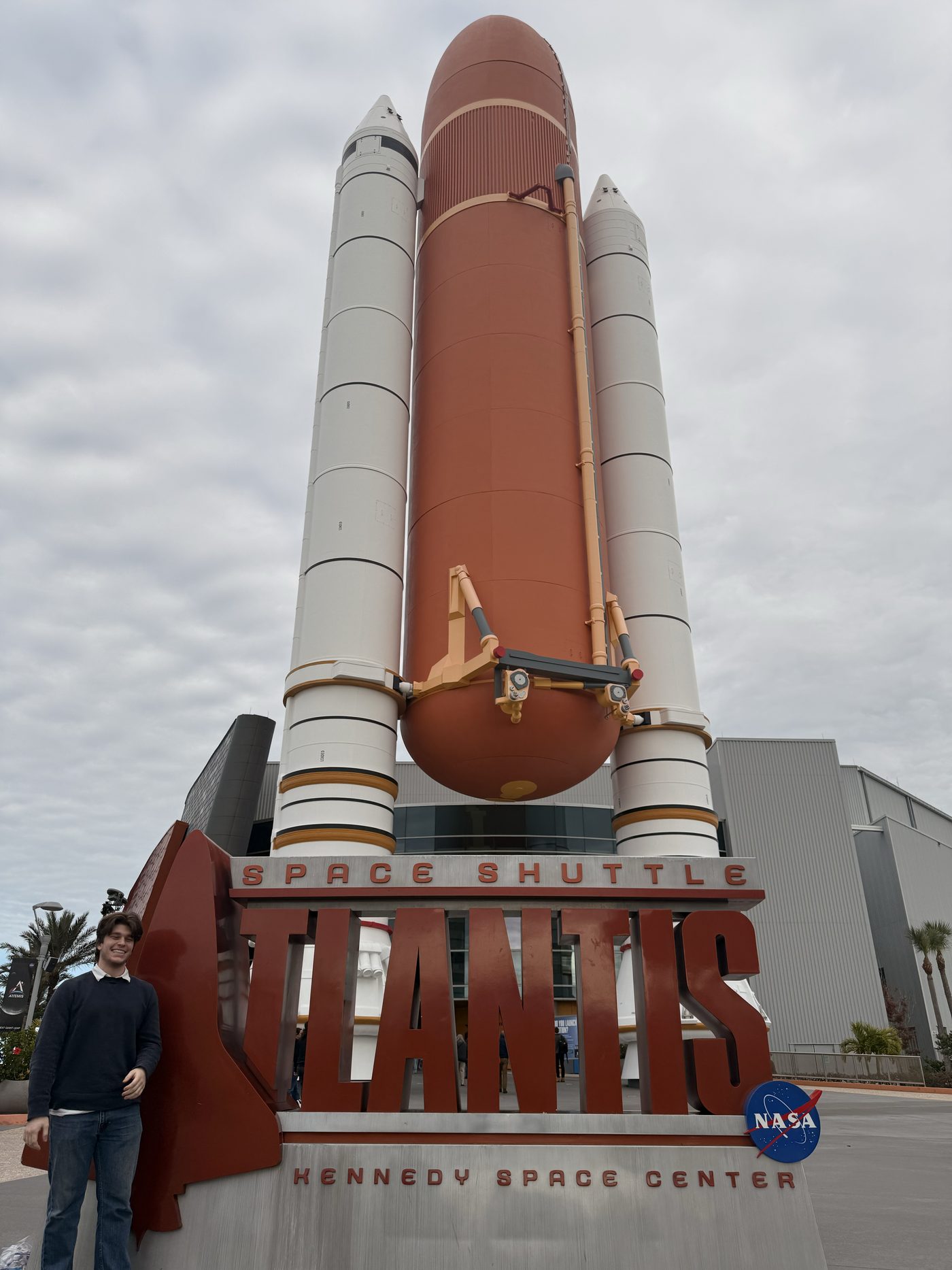 Benjamin Price standing beside the Space Shuttle Atlantis sign and stack model at Kennedy Space Center.